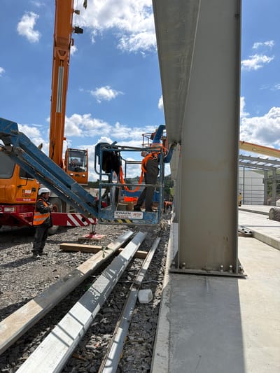 In Jodłownik, Poland, a construction site is visible under a partly cloudy sky. A large orange crane is present, with its boom extended upwards. A man wearing a hard hat and reflective vest stands near the base of the crane, holding a smartphone. A man in a harness is working in a cherry picker, appearing to be attaching a large metal beam to a building's structural frame. Several other workers are visible in the background, engaged in construction activities. Various construction materials and equipment, including metal beams and a large vehicle tire, are scattered across the gravel and concrete ground. The scene depicts active construction work on a building.