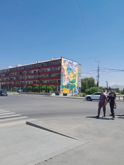In Artashat, Armenia, a bright, sunny day is captured, featuring a prominent red and white apartment building adorned with a vibrant mural on its side. The mural depicts a stylized landscape with mountains, trees, and figures. In the foreground, a pedestrian crossing is visible on a grey asphalt road. Several people are crossing the street, including a woman in a brown outfit, a boy in a black t-shirt, and another person whose features are obscured by the former two. A white car is stopped at a traffic light, and a black van is driving on the left side of the road. Trees and manicured bushes line the sidewalk in front of the apartment building. Power lines and streetlights are visible against the clear blue sky. The overall scene depicts a typical urban environment on a clear day.