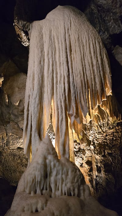 This media file is a close-up, landscape photograph of cave formations, likely stalactites and stalagmites, within a cave. The formations are predominantly white and cream-colored, with some appearing smooth and others more textured. Light, likely artificial, illuminates the formations from behind and within, highlighting their layered structures and creating a warm glow in some areas. The surrounding cave walls and ceiling are visible in the background, appearing dark, rough, and rocky, with patches of light reflecting off their surfaces. The image is shot from a low angle, looking upwards at the formations. There is no text visible in the image. The setting suggests a natural, underground environment. The texture and patterns of the mineral deposits are the primary focus, showcasing the intricate beauty of these geological formations. The lighting creates a dramatic and somewhat mysterious atmosphere. The location is identified as Vavřinec, Czechia, implying a specific geological site known for its cave systems.