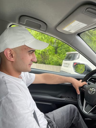 A man in a white baseball cap and a white t-shirt is inside a car, holding the steering wheel of what appears to be a Toyota. He is looking out the window. Outside the car, reflected in the side mirror, is the back of a white vehicle. Green foliage is visible in the background. A sun visor is flipped down on the driver's side. The location, based on the prompt, is Changilari, Georgia.