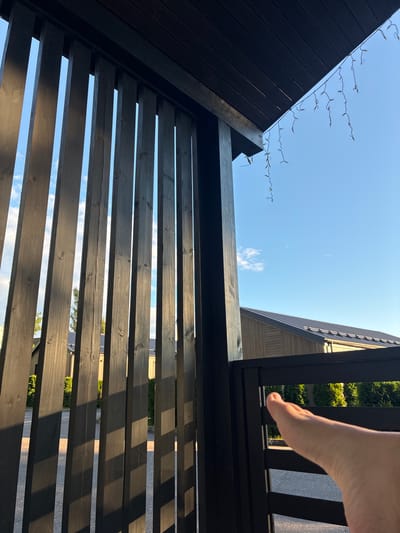 A low-angle shot captures a wooden structure with horizontal slats, possibly a fence or pergola, against a bright blue sky. The wood is dark, likely painted or stained, and shows visible grain and knots. Sunlight casts shadows through the slats, creating a striped pattern on the ground and the structure itself.

In the upper right quadrant of the image, a person's bare foot is visible, with the toes pointing towards the sky. The skin is lightly tanned, and the foot appears relaxed. This suggests a casual outdoor setting.

Along the top edge of the dark wooden structure on the left, a string of small, clear lights is draped. These lights are delicate and appear to be for decoration, possibly for an evening event.

In the background, beyond the wooden slats and against the sky, glimpses of a building with a sloping roof and vertical siding can be seen. Green foliage, likely trees or bushes, is also visible behind the fence.

The overall lighting and clear blue sky indicate it is daytime, likely late afternoon or early evening due to the distinct shadows. The scene appears to be an outdoor residential or recreational area. There is no visible text in the image. The setting is Peresvet, Russia.