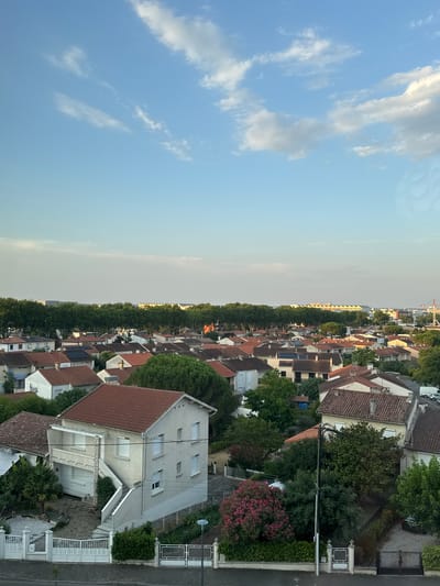 The image depicts a suburban residential area in Colomiers, France, on a clear day with scattered clouds. The scene is an aerial view looking down on a collection of houses with red-tiled roofs. The architecture suggests a typical European suburban neighborhood.
In the foreground, a light-colored house with a red roof is prominently featured. It has multiple windows and a balcony on the upper level. A staircase leads up to the entrance. The house is surrounded by greenery, including trees and bushes. A white metal fence and gate are visible in front of the property, along with a paved driveway.
Further back, more houses with red roofs are densely packed, interspersed with trees. A row of mature trees forms a green line across the middle ground, separating the immediate neighborhood from structures in the distance.
In the background, larger buildings, possibly commercial or industrial, are visible behind the tree line. A red construction crane is also present in the far distance, suggesting ongoing development or infrastructure projects. The sky is blue with streaks of white clouds, indicating fair weather. The overall impression is one of a peaceful, well-established residential district within a larger urban or peri-urban landscape. No people are visible in the image.
