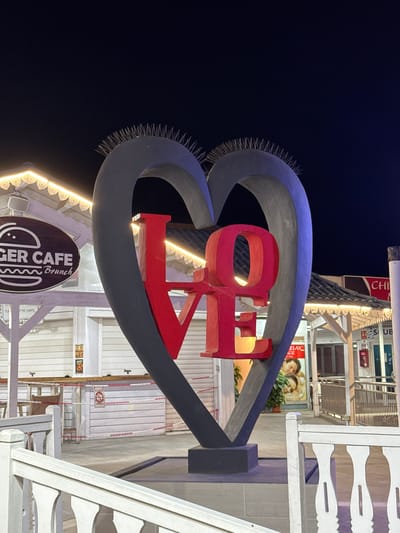This is a vertical, nighttime shot of a large, stylized heart sculpture with red letters spelling out "LOVE" inside. The sculpture is illuminated from below with warm yellow lights, creating a glowing effect. The heart structure itself is dark grey with a bright blue outer rim and has numerous thin, pointed metal spikes protruding from its outer edge, giving it a somewhat aggressive or protective appearance.
In the background, there are glimpses of a building with various signs and structures. One sign is partially visible on the left with the text "CHI". Another sign on the right reads "SUB". Further back, there's a poster featuring a couple, with the text "RELAX" and "AGE" visible. The overall scene suggests a commercial or entertainment area, possibly a boardwalk or a shopping street, given the signs and the illuminated structures. The presence of the "GER CAFE Brunch" sign at the bottom of the image provides a more specific clue about the location's offerings.
The lighting suggests it is evening or nighttime. The dark sky in the upper left corner confirms this. The mood is festive and romantic, implied by the heart shape and the word "LOVE". The presence of architectural elements like white railings and wooden paneling hints at a seaside or resort location. The image is framed to focus on the "LOVE" sculpture, with the background elements providing context without being the primary subject.
