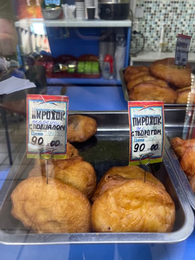 This image shows a close-up view of a display case filled with golden-brown, fried pastries. The pastries appear to be some kind of sweet buns or pies. Two price tags are visible, each attached to a pastry with a metal loop. The tags are in Russian and indicate the price is 90 rubles. One tag also says "жареный" (fried) and "с творогом" (with tvorog/cottage cheese), suggesting one type of filling. The other tag says "жарени" (fried) and "сподиалом" which is a less clear inscription but might refer to another filling or preparation method. The display case itself is made of metal and glass, with a blue background visible behind the pastries. The lighting suggests an indoor setting, possibly a bakery or a food stall. The overall impression is one of freshly prepared, tempting baked goods.