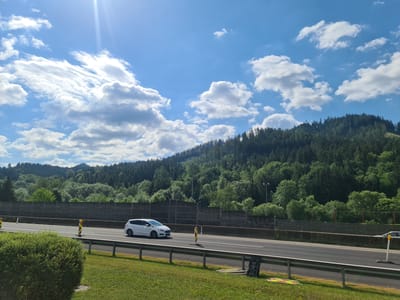 The image depicts a highway scene in Sankt Marein im Mürztal, Austria, under a bright, partly cloudy sky. A white car is traveling on the highway, which is bordered by a noise barrier and a grassy embankment. The background is dominated by a dense, green forest covering a hillside. Several traffic cones and construction materials are visible on the side of the road. The sun is shining brightly, casting a glare on the left side of the image.