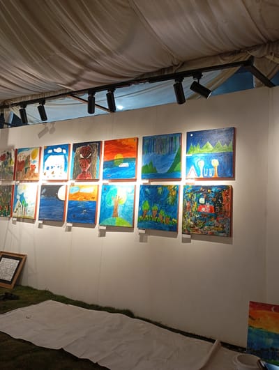 A wall display of children's paintings is showcased at an outdoor exhibition, possibly part of an event or festival in Meunasah Papeun, Indonesia. The paintings, arranged in two rows, feature vibrant colors and simple, childlike imagery, depicting scenes such as landscapes, buildings, and a portrait of Spiderman. Some paintings have labels affixed to the wall below them.

The exhibition area is covered by a white tent, with black track lighting illuminating the artwork. In front of the wall, a patch of green grass is visible, with a white sheet spread out on it, perhaps for laying out more artwork or for visitor seating. To the left of the display, a framed piece of artwork with handwritten text leans against the wall. The overall atmosphere suggests a community art event.