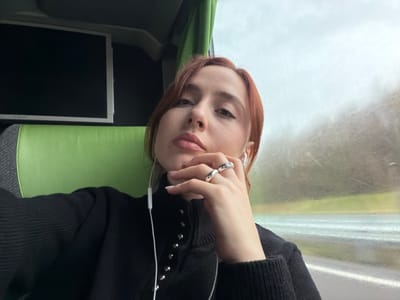 A red-haired individual is depicted taking a selfie inside a vehicle, identified as being in Courtisols, France. The person wears a black, high-collared ribbed sweater with a line of silver-colored buttons extending vertically from the collar. White wired earphones are visible, with the left earbud in place. The individual's left hand, adorned with two silver rings (one plain, one with a clear square stone), is positioned near the chin. The person's gaze is directed towards the camera. To the left, a bright green and grey upholstered seat is visible, unoccupied, with a dark, unlit monitor screen mounted above it. To the right, a window reveals a blurred, overcast outdoor landscape, suggestive of motion, with faint indications of trees, a road, and possibly rain on the glass. The interior lighting is consistent with ambient daylight filtering through the window.