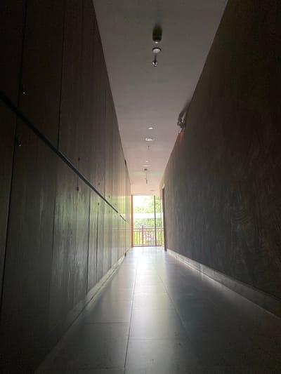 The image depicts a long, dimly lit hallway. The left wall is lined with vertical wooden panels, while the right wall has a darker, textured finish. The ceiling is white and features several recessed lights and what appear to be smoke detectors. A handrail runs along the left wall. The hallway opens to a brightly lit area at the end, revealing an outdoor space with a railing and foliage. The floor is made of large, light-colored tiles that reflect the light. The scene is devoid of any people or discernible actions. The location is described as being in the city of Pung Luong, Vietnam.