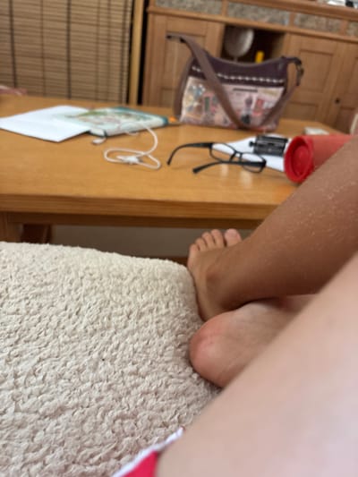 The media file shows a close-up, low-angle shot of a person's legs and feet. The person's left leg, covered in fine hairs, is positioned in the foreground, while their right foot is visible, resting on a textured, off-white surface, possibly a rug or blanket. The right foot is bare and appears to be a child's. The background is blurred but suggests an indoor setting. To the left of the legs, on a wooden surface, are several objects including a small brown handbag with a patterned insert and a strap, a pair of black-rimmed glasses, and a white cord object that resembles an earbud cord. There is also a red cylindrical object, possibly a container, and a piece of white paper with some colorful graphics. The lighting suggests it might be daytime, possibly indoors. The scene is static, with no discernible activity or strong emotional cues. The location context provided is Benissa, Spain, but this is not directly evident from the visual information in the image.
