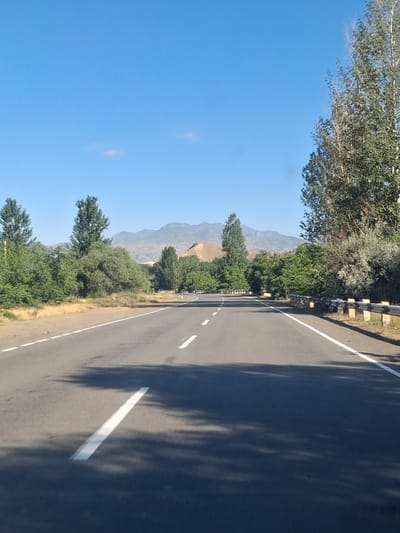 The image captures a straight, asphalt road with white lane markings, traveling through a rural landscape in the vicinity of Malishka, Armenia. The road is bordered by a shoulder on the left and a guardrail on the right. Lush green trees line both sides of the road, with a dense cluster on the left and a tall tree on the right. In the background, rolling hills and mountains are visible under a clear blue sky. A few scattered clouds are present in the sky. The scene appears to be during daylight, with shadows cast on the road, indicating the sun is at an angle. There are no people or vehicles clearly visible on the road, suggesting a quiet, undeveloped area.