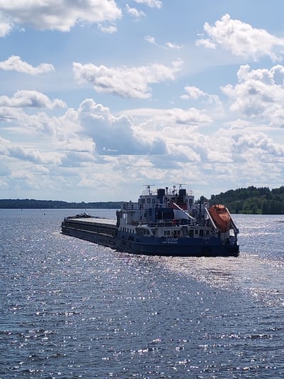 A barge is seen on a body of water, under a blue sky with white clouds. The barge is comprised of a dark hull, and a multileveled structure. A bright orange lifeboat is suspended on the back of the barge. The water surface reflects the sunlight, creating a shimmering effect. The background consists of a distant treeline on the horizon. Text on the hull reads "CECKAP," and "САНКТ ПЕТЕРБУРГ." The location is likely near Gorka, Russia.