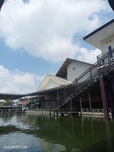 This image, captured on 2025/08/23 at 12:06, presents an outdoor scene in Palasari, Indonesia. The view is from a low angle, looking across a calm, dark green body of water that occupies the foreground and lower mid-ground, reflecting the sky and adjacent structures. The sky overhead is a light blue with scattered white cumulus clouds.

Architectural structures, likely part of a restaurant or resort complex, are visible across the mid-ground, built partially over or directly next to the water. On the far right, a light-colored building with a dark eave features a prominent dark metal staircase with mesh railings descending towards the water level. Adjacent to this, a multi-story building with a light-colored gable roof and lighter walls is visible. Below this, a section of the building features horizontal wooden or bamboo-like slats. Further to the left, a long, open-sided covered platform extends over the water, supported by pilings. This platform has a light-colored, possibly translucent, roof and appears to be a dining area with tables and chairs visible. Several orange buoyancy aids are hanging from the structure near this area. Distant structures with corrugated roofs and sparse foliage are visible in the background on the far left. Potted plants line the edge of some of the structures at water level. No individuals are present or engaged in any discernible actions within the frame.