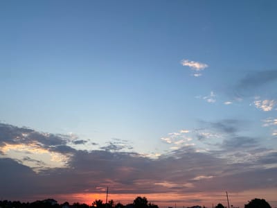 A wide-angle, full-frame, eye-level photograph captures a serene dusk sky over Kurtovo Konare, Bulgaria. The sky transitions from a pale, clear blue at the top to shades of pink and orange near the horizon, illuminated by the setting sun. Scattered clouds, some tinged with golden light, drift across the sky. Silhouetted against the vibrant sunset are the rooftops of houses and the dark shapes of trees, indicating a residential area. The overall impression is one of peaceful twilight.