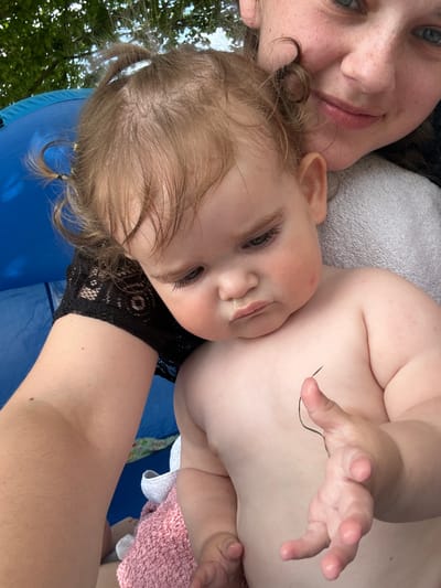 This is a close-up, vertically oriented selfie taken outdoors, likely during the daytime given the natural lighting and visible greenery. The primary subjects are a woman and a baby. The woman is in the foreground, her face partially visible in the top left corner, with green eyes and freckles. She is smiling. The baby is positioned next to her, facing the camera, with light brown hair. The baby's face is also in focus, and appears to be looking towards the camera with a somewhat relaxed expression, with a bit of liquid around its mouth. The baby is wearing a black, lace-trimmed garment around its neck and upper chest.

The setting appears to be outdoors with trees visible in the background, suggesting a natural environment. The subjects are resting on a blue inflatable surface, possibly a pool or a play mat. A light grey towel is visible behind the woman's head, and a pink towel is seen near the baby. The overall mood appears to be one of shared intimacy and enjoyment between the woman and the baby. There is no visible text in the image. The location context provided is Giessen, Germany.
