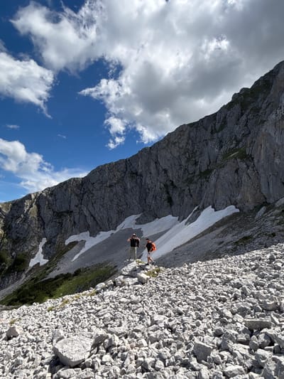 The image depicts a rocky, mountainous terrain in Pašina voda, Montenegro. Two individuals, likely hikers, are visible in the mid-ground. One person, wearing a dark shirt and light-colored shorts, stands facing the camera with their right arm raised. The second person, wearing an orange backpack and shorts, is further to the right and appears to be walking away from the camera. The landscape is characterized by a steep, grey rock face dominating the background, with patches of snow and green vegetation visible along its base. The foreground is covered in a dense scattering of grey rocks and stones. The sky above is a vibrant blue with scattered white and grey clouds.