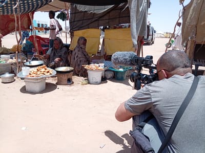 The image captures an outdoor market scene in Goshabi, Sudan, where daily activities are being documented by a film crew. In the foreground, a man, seen from behind, is crouched down, intently operating a professional video camera. The camera is fitted with a large furry wind-muff on its microphone, and the man wears glasses, a grey t-shirt, and a dark shoulder bag.

His camera is pointed towards a makeshift food stall set up under a canopy of various fabrics, including a patterned blue and white cloth and dark tarpaulins, supported by wooden poles. Two women are seated on the sandy ground, actively preparing food. The woman on the left, dressed in a patterned garment and head covering, is stirring a large pan with a ladle. Next to her is a large tray filled with golden-brown fried food, resembling fritters or donuts, and a traditional clay or mud oven. The second woman, further back and partially obscured, is also seated, wearing a similar head covering. White and green plastic buckets, metal pots, and various utensils are arranged around their cooking area. One white bucket displays visible text in Arabic script, alongside the English word "Organisation," suggesting it might be from a humanitarian or development group.

In the mid-distance, a young man in a pink t-shirt stands near the cooking area. Beyond the immediate stall, the scene opens to a dusty, arid landscape under a clear, bright sky. Several people are visible in the background, including a man in traditional white clothing pushing a wheelbarrow, and a white SUV. A light blue building can be seen in the far distance, providing a contrasting, more established element to the rustic surroundings.

The bright sunlight and strong shadows indicate it is daytime, likely midday or early afternoon. The overall impression is one of a bustling, yet routine, outdoor marketplace in an arid region, with the film crew observing and recording local life.