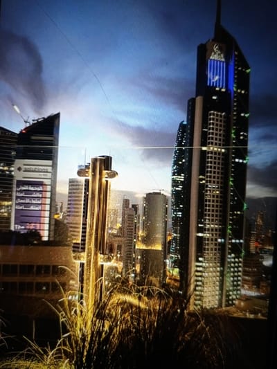 The image shows a cityscape at dusk, with several skyscrapers illuminated by lights. In the foreground, tall, dry grasses are silhouetted against the city lights. The sky is a deep blue, transitioning to lighter shades near the horizon, suggesting the end of the day.
The most prominent building in the center is a very tall skyscraper with a dark facade. It has a distinctive logo at the top and vertical blue lights running down a section of its side. To its left is another skyscraper with a more angular design. A large sign on this building displays "solutions" in white text on a dark background. Below this, a purple banner has several lines of Arabic text, followed by "The 471".
Other buildings of varying heights and designs are visible in the background, some with grid-like window patterns and others with scattered lights. The overall impression is one of a modern, vibrant urban environment at twilight. The presence of the dry grass in the foreground suggests an outdoor viewpoint, possibly from a balcony or a higher elevation overlooking the city. There is no discernible activity or event taking place; it is a static scene of urban architecture. The lighting and the time of day create a somewhat dramatic and sophisticated atmosphere.