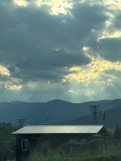 The image depicts a landscape scene with a building in the foreground, mountain ranges in the middle ground, and a dramatic sky with sunbeams breaking through clouds in the background. The building is dark with a corrugated metal roof. It has at least one window visible. In the distance, there are two towers, one with a Bulgarian flag. The mountains are layered, fading into a hazy blue in the distance. The sky is filled with dark, heavy clouds interspersed with bright areas where sunlight is piercing through, creating visible sunbeams. The overall atmosphere is moody and dramatic. The location is identified as the city of Pavelsko, Bulgaria.