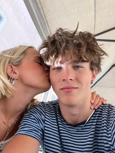 A close-up, selfie-style photograph features two individuals, a blonde female and a brown-haired male, in an outdoor setting beneath a beige shade umbrella. The female, positioned on the left side of the frame, has shoulder-length blonde hair, light complexion, and her eyes closed while kissing the male's right cheek. She wears multiple silver earrings, including one with three small white pearls, and several silver necklaces, one adorned with small green beads. Her right hand, exhibiting multiple rings, rests on the male's left shoulder and neck. The male, central in the image, has messy, light brown hair, blue eyes, and looks directly at the camera with a neutral expression. A bright, focused light spot is visible on his forehead. He wears a short-sleeved shirt with horizontal light blue and dark blue stripes, a silver hoop earring in his left ear, and a flat silver chain necklace. In the upper left background, a light-colored building with a dark-framed window or ventilation unit is partially visible. While the scene is located within the city of Foligno, Italy, no specific landmarks or discernible features in the image permit the identification of an exact address or precise location within the city.