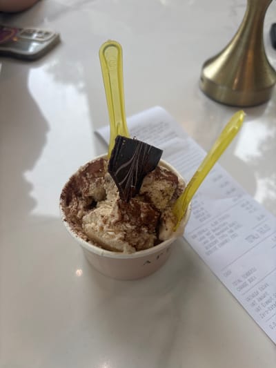 A close-up, eye-level shot captures a cup of gelato with two yellow plastic spoons and a piece of dark chocolate. The gelato is creamy beige, dusted with cocoa powder. The cup is pinkish-white with the word "AP" visible on the side. The scene is set on a white marble surface, with a receipt and a golden ornament visible in the background. The image appears to have been taken at the SM Mall of Asia in Pasay City, Philippines, specifically within a dessert shop like a gelato parlor.