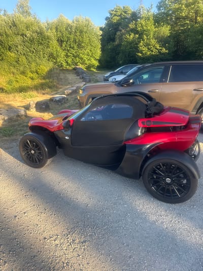 A low-angle, full shot captures a red and black three-wheeled sports car parked on a gravel surface. The car is positioned on the right side of the frame, facing left. It has a sleek, aerodynamic design with a black fabric roof. Behind the sports car, the rear section of a tan SUV is visible, indicating a parking area. The background consists of a dense line of green trees and vegetation, illuminated by warm, late-day sunlight. A rough stone wall runs along the base of the vegetation. The sky is clear and blue. The scene appears to be an outdoor parking lot adjacent to a natural, wooded area. The car has the number "115" in a red circle on its rear panel.