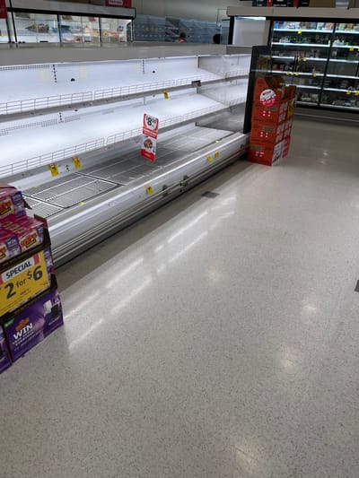 The image shows a grocery store aisle in Middle Park, Australia (latitude: -27.556, longitude: 152.923). The aisle is empty except for a stack of cardboard boxes and a few packages of a product labeled "SPECIAL 2 for $6". The aisle is lit by overhead fluorescent lights. The floor is a light gray speckled tile. The shelves are metal and have a white finish. The shelves are empty, except for a few yellow price tags. The aisle is lined with white walls. The shelves are surrounded by a metal frame. The floor is covered with a grey and white speckled pattern tile. The ceiling is out of the frame.