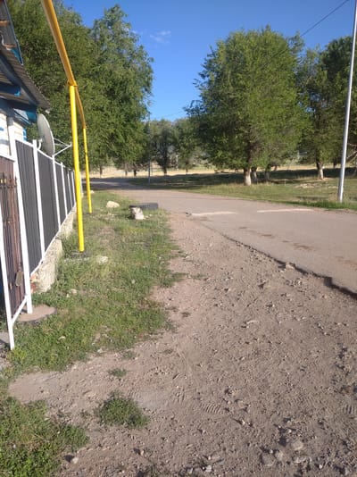 The image captures a roadside scene in Kygali, Kazakhstan, under a clear blue sky. In the foreground, a dirt and gravel path runs parallel to a paved road. To the left, a low, white-brick building with a corrugated metal roof is partially visible, bordered by a black and white metal fence. A satellite dish is mounted on the fence. A bright yellow gas pipe runs vertically along the fence line, supported by metal posts. Beyond the road, a grassy area is dotted with mature trees, their trunks appearing light-colored. The road itself shows signs of wear, with cracks and uneven surfaces. The overall environment suggests a rural or semi-urban setting on a sunny day.