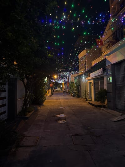The image depicts an alleyway in Nam Nha Trang, Vietnam, illuminated by strings of multicolored lights spanning overhead. A Vietnamese flag hangs from a building on the left. Several people are walking through the alley. Buildings line both sides of the alley, with a mix of residential and commercial structures. The pavement appears uneven and aged. Vegetation, including trees and bushes, is visible along the sides. A utility access panel is visible on the right.