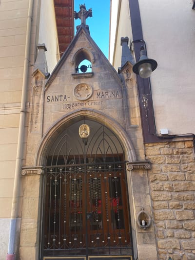 A forensic description of the image reveals the entrance of a stone-built structure identified as the Santa Marina chapel or hermitage, located in Zarautz, Spain. The facade features a pointed archway leading to a brown wooden door, obscured by an ornate black wrought-iron gate with gold-colored decorative elements. Above the gate, a golden, circular emblem depicting a figure (possibly a bust) is suspended.

Carved into the stone lintel above the arch are the words "SANTA-MARINA" and below it, "1932GARen URTEAN," indicating "in the year 1932." Above these inscriptions, a quatrefoil-like stone carving is centered, with a small bell suspended in an open archway above it. The structure culminates in a stone cross at its apex, flanked by decorative pinnacles.

To the left of the entrance is a smooth, light-colored building with a vertical grey downspout. To the right, a building with a beige stucco upper section and a rough-hewn stone lower section is visible. A black, conical street lamp with a clear glass globe extends from the right building's facade. Below this lamp, a small, shell-shaped stone niche is affixed to the stone wall, containing a silver-colored beverage can. The upper portion of a wooden balcony is visible between the buildings, against a clear blue sky. No individuals are present in the scene.