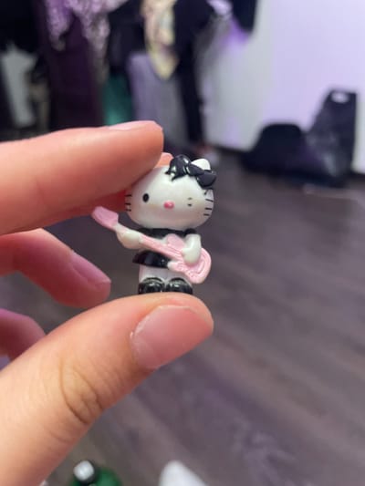 A close-up, shallow depth of field shot shows a person's fingers holding a miniature Hello Kitty figurine. The figurine is white with a black bow and black dress, and it is holding a pink toy guitar. The background is blurred and shows what appears to be a wooden floor and some indistinct objects in the distance. There is no discernible location within Borisova Griva, Russia.