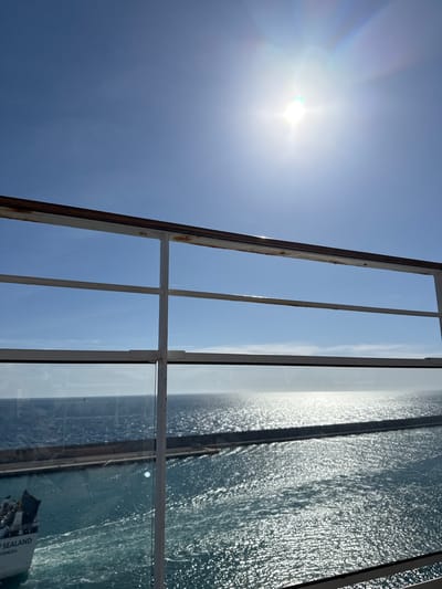 The image captures a bright, sunny daytime scene overlooking a harbor or port in Civitavecchia, Italy. The vantage point is elevated, likely from a ship's deck or a building, framed by a white metal railing with a worn, rusted brown top bar and a clear glass pane in the foreground.

The sky occupies the upper portion of the frame, a clear and vibrant blue, with the sun positioned in the upper right quadrant, emitting a strong lens flare and distinct sun rays.

Below the horizon, the sea extends, reflecting the intense sunlight with a glittering, shimmering surface, particularly towards the right. In the midground, a dark, linear breakwater or pier structure runs horizontally across the water.

To the lower left, partially visible through the glass pane and behind the railing, is the stern of a white ship. The words "SEALAND" and "VENEZIA" are legible on its hull, indicating the vessel's name or operator and possibly its port of origin or destination. The ship is moving, creating a white wake behind it in the turquoise water, suggesting it is either departing or arriving within the harbor area defined by the breakwater. No individuals are discernible in the scene.