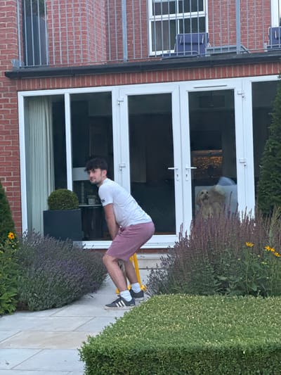 The scene depicts the exterior of a two-story brick residence in Bubwith, United Kingdom, featuring a paved patio area and landscaped garden. A young adult male, with dark, curly hair, is positioned in a crouched stance on the patio, facing partially away from the camera. He wears a white t-shirt, pink shorts, white socks, and dark espadrille-style shoes with white stripes. He holds a vertical yellow object, consistent with part of a pogo stick, between his legs. His head is turned to his left, looking towards the right side of the frame.
Behind the male are large, white-framed glass bi-fold or French doors leading into the residence's interior. Through the glass, a light brown dog with floppy ears is visible, standing and looking towards the camera. Inside the residence, a dark planter with a spherical green plant is visible to the left, and a dark light fixture hangs from the ceiling.
Above the ground floor, the residence features red brickwork with a first-floor balcony equipped with a dark metal railing and vertical bars. Two blue outdoor chairs are visible on this balcony. The foreground of the image includes a manicured green hedge. To the left of the male, a bush with purple flowers (likely lavender) borders the patio, while to his right, another bush with darker stalks and yellow flowers is present. A tall, narrow evergreen tree is partially visible on both the left and right sides of the frame. The lighting suggests late afternoon or early evening.