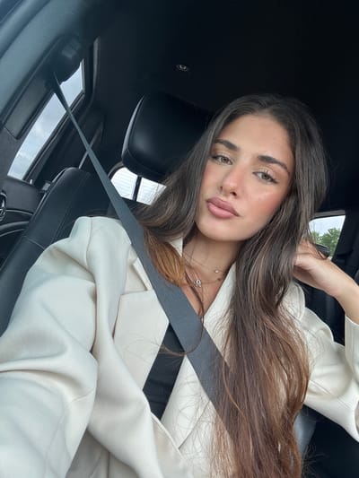 A young adult female is in the driver's seat of a car. She wears a beige blazer, a seatbelt, and a necklace. The location is presumed to be North Miami, Florida, United States.