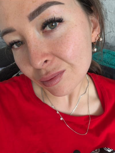 A close-up, waist-up selfie of a woman with light skin, freckles, and striking green eyes. She has full lips with a subtle pink lip gloss and dark, well-defined eyebrows. Her eyelashes are long and dark, appearing to be extensions. She is wearing a bright red t-shirt and a delicate silver necklace with small diamond-like charms. Her left ear is adorned with a gold hoop earring featuring a star shape and a dangling charm. Her hair is brown and partially visible. The background is a neutral gray textured wall, with hints of green and black fabric to the right. The image is taken indoors, and there is no discernible indication of location within the city of Япрыково, Russia.