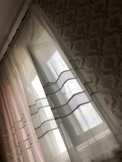 The image depicts a close-up view of a window with curtains. Two layers of curtains are visible: a pinkish-toned one on the left and a sheer white one with horizontal striped detailing on the right. The white curtain is partially drawn, allowing light to filter through from the window behind it. The window itself appears to be a standard residential window, likely in an apartment or house. In the background, the wall is covered with patterned wallpaper. The overall lighting suggests it is daytime, with the light casting shadows and highlighting the textures of the curtains and wallpaper. The scene is indoors, focusing on the window treatment and the texture of the wall. There are no people or specific actions depicted. The location in Calfa, Moldova, is not visually ascertainable from the image itself.