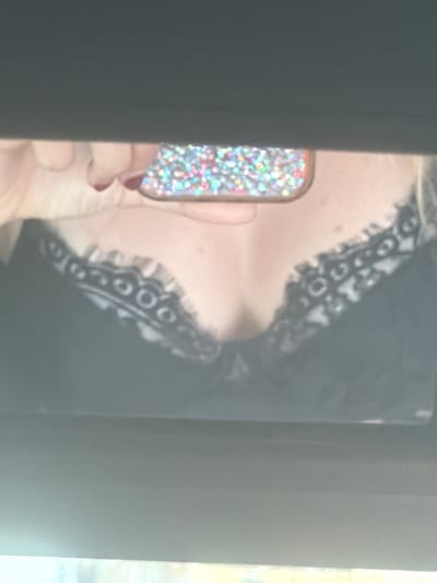 The image shows a close-up of a person's chest wearing a black lace bra. A hand, with painted fingernails, is holding a phone with a sparkly, multi-colored case. The phone is reflecting light, and the image appears to be a reflection in a mirror. The lighting is dim, and the background is out of focus. There is no discernible information about the location in San Vicente Del Raspeig/Sant Vicent Del Raspeig, Spain.