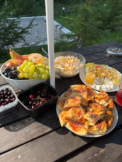 This is an overhead, slightly angled medium shot of a picnic spread on a dark wooden table outdoors, likely in Kuliki, Russia. The table is laden with various food items and a white pole, possibly part of an umbrella or tent, bisects the image vertically.
In the foreground to the left are two plastic containers. The container on the left holds dark berries, possibly blueberries or blackcurrants. The container to the right holds dark red cherries.
To the right of the containers is a large white bowl overflowing with a variety of fruits, including green and dark grapes, sliced pears, and red apples.
Behind the fruit bowl is a glass bowl filled with a creamy salad containing corn and other ingredients.
To the right of the salad is a white plate with an assortment of cheese, including wedges of brie and cubes of yellow cheese, accompanied by what appears to be small nuts in a shot glass of orange juice. There are also some pieces of what looks like grapefruit or pomelo on the plate, garnished with small toothpicks.
Further to the right, and closer to the foreground, is a white plate piled high with sliced baked goods. These appear to be a mix of savory items, including what looks like small pizzas with cheese and ham, and sweet items, such as apple pie or tart slices with browned crust.
A glass of red liquid, likely juice or wine, is visible on the far right edge of the table.
The background shows lush green trees and some structures, suggesting a natural setting. The lighting indicates it is daytime, with sunlight casting shadows on the wooden table. There are no people visible in the image.