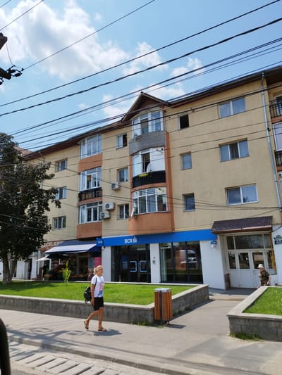 In Bragadiru, Romania, on a sunny day, a woman walks along a sidewalk in front of a commercial building. The building has a light beige facade with orange accents and multiple windows. A blue awning extends from a storefront, and the BCR bank logo is visible on the building. A large tree with dark foliage is on the left side of the image. Overhead, numerous power lines crisscross the sky, which is partly cloudy. A woman in a white t-shirt and black shorts walks from left to right, carrying a black backpack. To her right, a person wearing a camouflage shirt and a white hat stands near an entrance. The scene depicts an urban street environment with commercial establishments and residential apartments.