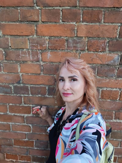 A woman with light pink, wavy hair stands in front of a brick wall. She is wearing a black top with lace trim and a colorful, abstract-patterned jacket in shades of pink, orange, grey, and blue. A light yellow backpack is visible on her right shoulder. Her right hand, with red nail polish, is resting on the brick wall. She is wearing a black ring on her index finger. The woman is looking towards the camera with a slight smile. The brick wall appears aged with some visible mortar. The scene is an outdoor urban setting.