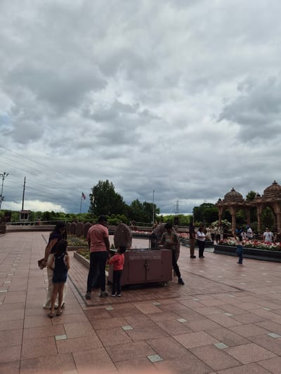 The image depicts the exterior of a religious site in Robbinsville Township, United States of America. The sky is overcast with heavy clouds. In the foreground, several people are gathered on a tiled plaza. A family, including two adults and two children, stand near a decorative metal box. To the right, another group of individuals are observing the surroundings, with some looking towards a building with ornate domes in the background. The environment features manicured flower beds and large stones. In the midground, an American flag is visible. Power lines and utility poles are also present in the background, suggesting an urban or suburban setting.