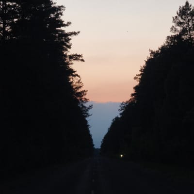 A dimly lit road stretches into the distance, flanked by silhouetted trees against a twilight sky. The road has a white line dividing lanes. A single light source, possibly headlights, is visible further down the road. The location appears to be Парэцкі сельскі Савет, Belarus, based on the presence of trees and a rural road. The time is dusk.