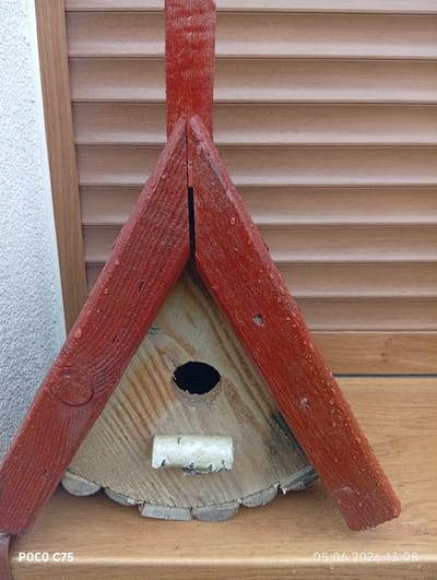 The image displays a rustic, handmade birdhouse positioned on a light wooden surface, likely a windowsill, in Kiełczów, Poland. The birdhouse features a prominent A-frame roof structure, painted a distinct reddish-brown, with numerous water droplets visible on its surface, indicating recent rain or dew. The main body of the birdhouse is crafted from natural, light-colored wood, possibly a cross-section of a log, exhibiting prominent wood grain patterns. It has a circular entrance hole and a small, light-colored perch made from a branch directly below it. The base of the birdhouse appears to be a thick, round slice of wood with decorative, scalloped edges.

In the background, a window is partially visible, covered by horizontal wooden blinds or shutters of a light brown hue. To the left, a textured, off-white wall, possibly stucco, can be seen. The wooden surface on which the birdhouse rests complements the warm tones of the background blinds.

No human subjects or explicit activity are depicted, but the water droplets on the birdhouse suggest it has recently been exposed to moisture. The visible text "POCO C75" is in the bottom left corner, indicating the device used. A timestamp "05.06.2026 13:08" is present in the bottom right corner, showing the photograph was taken in the early afternoon. The overall scene conveys a simple, outdoor domestic setting.