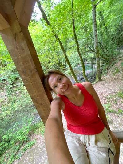 In this media file, a woman with blonde hair and fair skin is taking a selfie in a natural setting. She is wearing a red, ribbed tank top and light-colored shorts. Her right arm is extended, holding the camera, and her left hand rests on a wooden beam. The woman is looking at the camera with a slight smile.

The background reveals a lush, green forest with sunlight filtering through the leaves. Several trees with mossy trunks are visible, and a body of water, possibly a river or stream with a turquoise hue, can be seen in the distance. The ground is covered with dirt, leaves, and some small plants. The overall scene suggests a daytime outdoor activity in a woodland area. The woman appears relaxed and happy. There is no visible text in the image. The location context of Izmaylovka, Russia, further grounds the scene in a specific geographical place.
