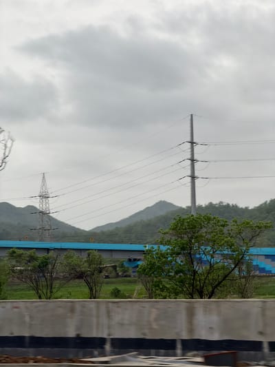 The image depicts a cloudy day in Saphale, India. In the foreground, a concrete barrier with black stripes runs horizontally across the frame. Behind the barrier, several trees with green foliage are visible. A long, blue and grey structure stretches across the mid-ground, with a bridge element visible within it. In the background, rolling green hills are partially obscured by a layer of mist or clouds. Power lines crisscross the scene, supported by tall, metal pylons. One pylon is a lattice tower, while another is a single, tall pole. The overall atmosphere is overcast and somewhat muted. No people or animals are visible in the scene.