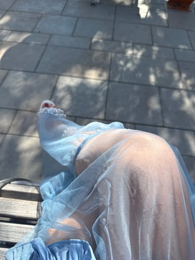 A close-up, high-angle shot focuses on a person's legs and feet, draped in a sheer, pale blue fabric, likely a dress or skirt, with subtle ruffles at the hem. The fabric is adorned with small, shiny embellishments, possibly pearls, around the ankle area. The person's bare legs are visible through the translucent material, with sunlight casting dappled shadows from unseen foliage onto the paved ground. The paving stones appear to be laid in a uniform pattern. A wooden bench is partially visible in the lower left corner, suggesting an outdoor seating area. The scene is set in a bright, sunlit environment. The specific location within Plainview, United States of America, is not identifiable from the visual information provided.