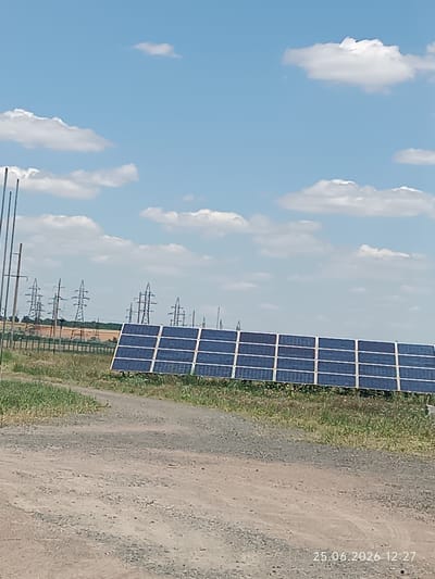 This media file depicts an outdoor scene with a clear blue sky dotted with fluffy white clouds. In the foreground, a gravel path leads towards a solar panel installation. The solar panels are arranged in multiple rows and are tilted towards the sky. Beyond the solar panels, a series of power lines and transmission towers stretch across the landscape, indicating a rural or industrial setting. The ground is covered with dry grass and some sparse vegetation. The overall impression is one of a bright, sunny day. The date "25.06.2026 12:27" is visible in the bottom right corner of the image, suggesting the time and date the photograph was taken. The location context is Artsyz, Ukraine. No people or animals are visible in the image. The activity taking place is the generation of solar energy.