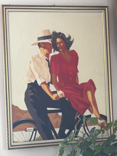 The media file is a framed painting depicting two people on a bicycle. The subjects appear to be a couple, with a woman in a red dress and a man in a white shirt and dark pants. The woman is leaning back, with her legs raised, as if embracing the man. The man is positioned in front of her, steering the bicycle. The style of the painting is illustrative, with bold colors and simplified forms. The background is a light, neutral color, which helps to highlight the figures and the bicycle.
The activity taking place is a romantic or playful moment on a bicycle, possibly a shared ride or a dance-like pose. The mood seems joyful and intimate, with the embrace and the dynamic posture suggesting a sense of movement and connection. There are no visible cues about the time of day, weather, or specific location within Uhersko, Ukraine. The framing suggests the artwork is displayed indoors, perhaps in a home or gallery. The painting is framed with a decorative border, adding to its presentation.