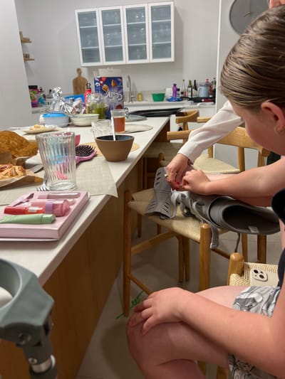 The scene takes place in a kitchen in Ramat HaKovesh, Israel. A young woman is sitting on a stool, facing the camera. Her right leg is in a gray orthopedic boot, and she is holding a part of it with her right hand. A woman is standing behind her, facing the camera, holding a pair of tongs. Both are wearing white clothing. The kitchen is in a state of disarray, with a variety of objects on the countertop, including a glass with a brown liquid, a glass with a pink liquid, a bowl, and a plate with pastries. There are also a variety of items on the shelf above the counter, including a box of cereal, a bag of food, and a variety of other household items. The floor is tiled in a light color.