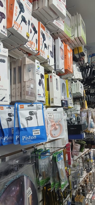 This image shows a retail store, likely in Akmachit, Uzbekistan, with numerous electronic accessories, primarily earphones, displayed on shelves and hanging racks. The products are packaged in boxes and plastic bags, with various brands and models visible. The overall impression is one of a well-stocked electronics shop. There are no people visible in the image. The environment appears to be indoors, under artificial lighting.