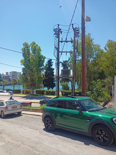 A green Mini Countryman is parked on a street. A silver Opel Astra is parked behind it. A red trash can is visible between the two cars. A utility pole with electrical equipment stands on the right side of the image. In the background, there is a body of water with buildings on the far bank, and trees line the waterfront. The sky is clear blue. The scene appears to be in a coastal urban area, possibly in Greece, given the style of buildings and the vegetation.